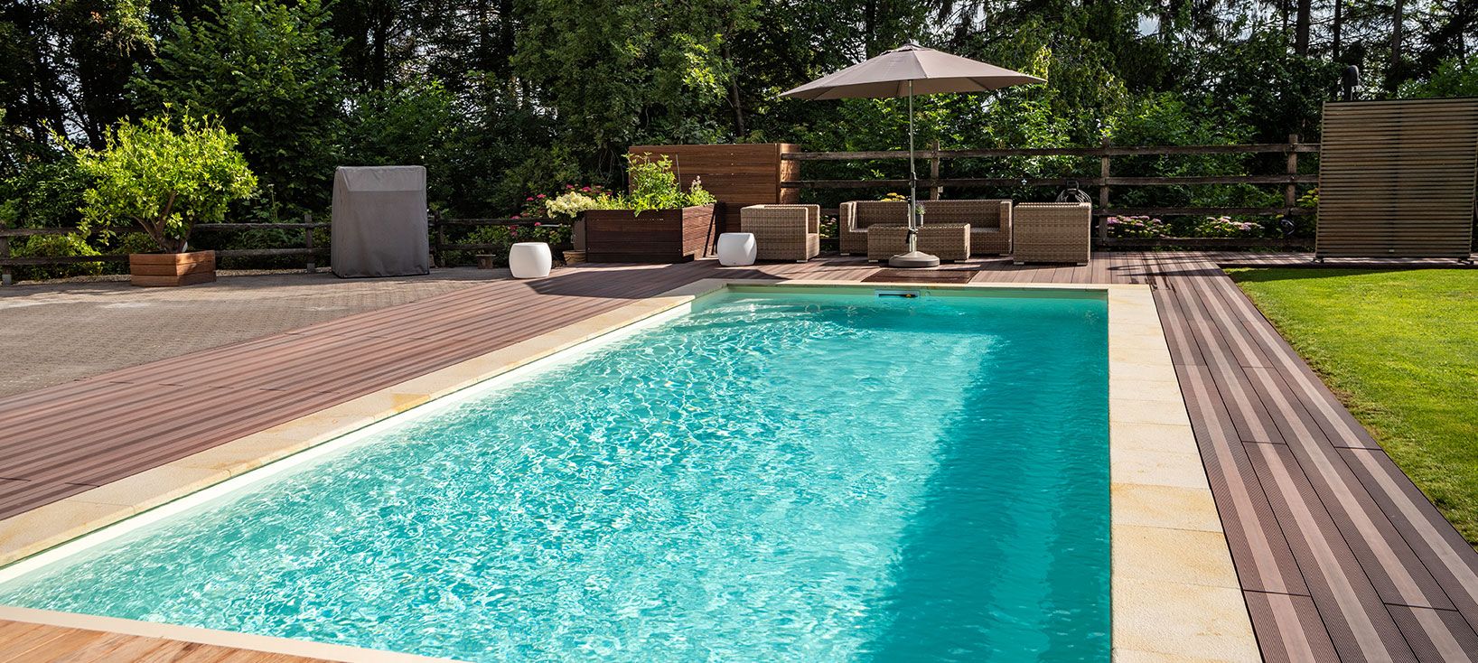 Rechteckiger Pool mit klarem Wasser, umgeben von Holzterrasse, Gartenmöbeln und grünem Rasen.